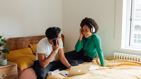 Friends sitting together on bed with laptop and headphones.
